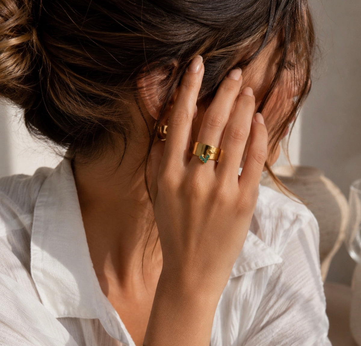 Bague Liza dorée avec petite breloque verte portée au doigt d'une femme en chemise blanche
