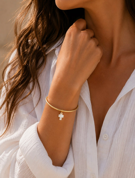 Bracelet jonc Victoire doré avec breloque croix blanche nacrée, porté au poignet d'une femme en chemise blanche